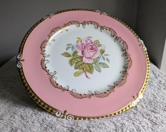 May include: A pink porcelain plate with a gold rim and a central floral design. The plate features a pink rose with green leaves and a decorative gold border. The plate is a decorative item.