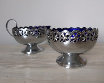 May include: Two silver-toned bowls with intricate cut-out designs around the rims. The bowls have a deep blue interior and sit on pedestal bases. One bowl has a handle. The bowls are likely for serving.