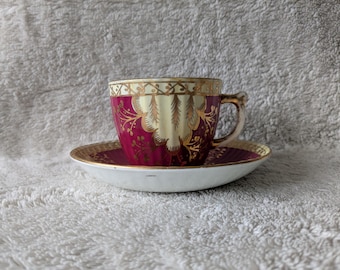 May include: A vintage teacup and saucer set. The cup and saucer are white with a burgundy and gold floral design. The cup has a gold handle and rim. The saucer has a matching gold rim and design. The set is likely porcelain.