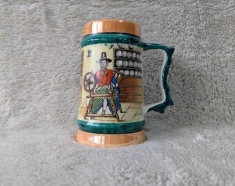May include: A ceramic beer stein with a handle, featuring a painted scene of a person using a spinning wheel. The stein has a green and orange trim, with a detailed illustration of a person in traditional clothing. The handle is a dark green color.
