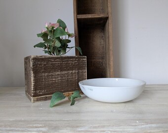 May include: A rustic wooden planter box holds a small plant with pink flowers and green leaves. A white ceramic bowl sits on a light-colored wooden surface, with a tall, weathered wooden shelf in the background.