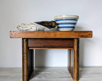 May include: A rustic wooden stool with a dark brown bottle, a white cloth, and a ceramic bowl with blue stripes. The stool has a rectangular top and a lower shelf, with four sturdy legs. The items are arranged on the stool.
