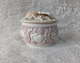 May include: A small, round trinket box with a lid, featuring a floral design on top. The box is cream-colored with a raised, detailed pattern of hearts, leaves, and flowers in a muted rose hue. The lid is a light beige color.