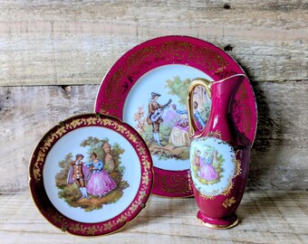 May include: A set of vintage porcelain tableware featuring a burgundy and gold color scheme. The set includes two plates and a pitcher, each adorned with romantic scenes and intricate gold detailing. The plates and pitcher are displayed on a wooden surface.
