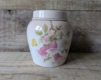 May include: A white ceramic jar with a lid, decorated with a floral pattern in pink, yellow, and green. The jar has a rounded shape and sits on a wooden surface. The floral design includes bell-shaped flowers and leaves.