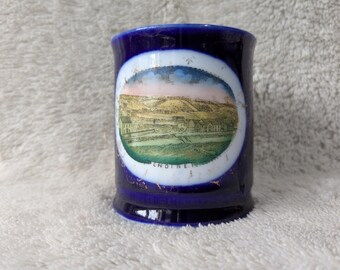 May include: A cobalt blue ceramic mug with a white oval design featuring a landscape scene. The scene depicts a village with rolling hills and the word "ENDINE" at the bottom. The mug has a flared base and a white interior.