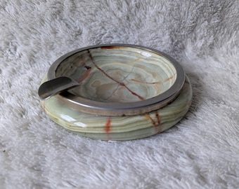 May include: A round ashtray made of green onyx with a silver-colored metal rim. The ashtray has a built-in rest for cigarettes. The onyx has natural veining in shades of green, brown, and red.