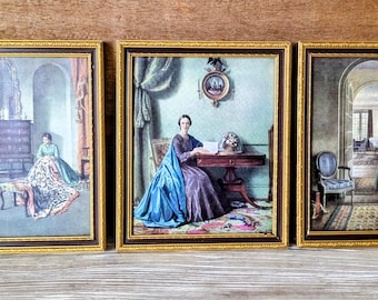 May include: Set of three framed vintage prints. Each print features a woman in a period dress in a room setting. The frames are gold-colored with a decorative edge. The prints have a muted color palette with shades of blue, green, and brown.