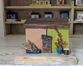 May include: A handcrafted wooden miniature house with a brown roof, pink and green walls, and a small blue planter. A small tree and a wooden stump are also present. The house sits on a wooden base, with other miniature houses in the background.