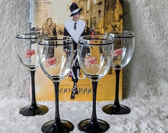 May include: Four clear wine glasses with black stems and rims. Each glass features a pink floral design. Behind the glasses is a book with the title "Exquisite" and the text "Harpers & Queen" and an image of a person.