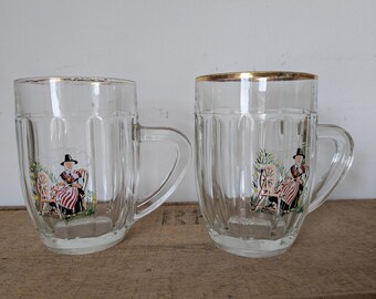 May include: Two clear glass beer steins with gold rims and handles. Each stein features a colorful illustration of a person in traditional clothing seated near a spinning wheel. The steins are on a wooden surface.