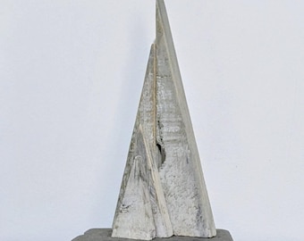 May include: A gray, abstract wooden sculpture with a triangular shape, mounted on a rectangular base. The sculpture has a rough, textured surface and a hole near the center. The artwork is displayed against a white wall.