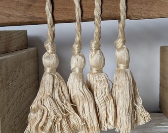 May include: Four beige tassels hanging from a wooden beam. Each tassel has a braided top and a full, fringed bottom. The tassels are a neutral color, suitable for various decor styles. The wooden beam is a light brown color.