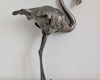 May include: A silver-colored flamingo sculpture. The bird has a long, curved neck and stands on two thin legs with webbed feet. The flamingo's feathers are detailed with textured patterns, and its beak is slightly curved.