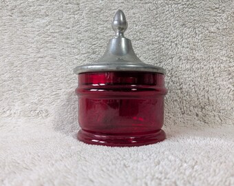 May include: A ruby red glass jar with a silver-colored metal lid. The jar has a cylindrical shape with horizontal ridges and a rounded base. The lid has a conical finial. The jar is likely for storage or display.