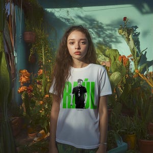 Könnte beinhalten: Ein weißes T-Shirt mit einer Grafik einer Person in einem schwarzen Trikot mit der Nummer 9, vor einem grünen Hintergrund mit dem Namen "RAÚL". Das T-Shirt wird von einer Person vor einer türkisfarbenen Wand und verschiedenen Topfpflanzen getragen.