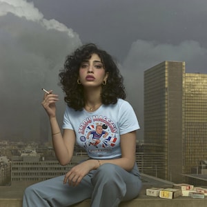 May include: A light blue t-shirt with a graphic of a soccer player and the text "Zizou Maestro". The person is wearing gold hoop earrings and a gold necklace. The background includes a cityscape and a cloudy sky.