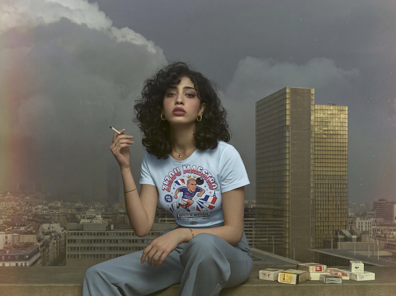 May include: A light blue t-shirt with a graphic of a soccer player and the text "Zizou Maestro". The person is wearing gold hoop earrings and a gold necklace. The background includes a cityscape and a cloudy sky.