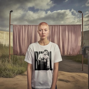 May include: A woman with a shaved head is wearing a white t-shirt with a black and white image of a boxer on the front. The shirt has the word "RIVAL" printed on it. The woman is standing in front of a pink curtain.