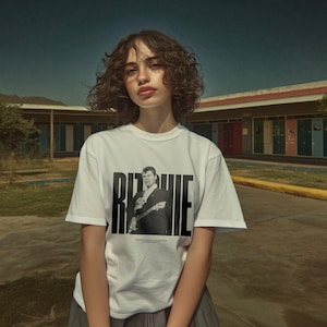 May include: A white t-shirt featuring a black and white image of a man with the name "Ritchie" printed in large, bold, black letters. The shirt is being worn by a person in front of a building with colorful doors.