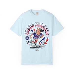 May include: Light blue t-shirt featuring a cartoon illustration of a soccer player kicking a ball. The design includes the text "Zizou Maestro" and "Artiste de Marseille" with French flag elements. The shirt is a vintage-style design.