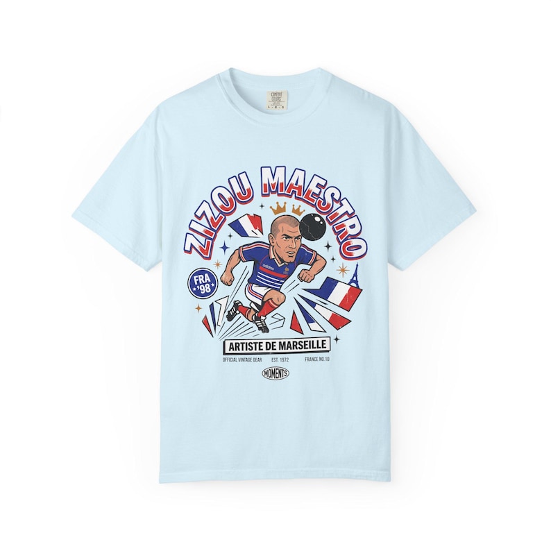 May include: Light blue t-shirt featuring a cartoon illustration of a soccer player kicking a ball. The design includes the text "Zizou Maestro" and "Artiste de Marseille" with French flag elements. The shirt is a vintage-style design.