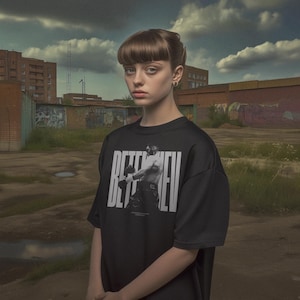 Puede incluir: Una persona joven lleva una camiseta negra con un gráfico blanco de un boxeador. La camiseta tiene el texto "BEETLEHEU" impreso. La persona está de pie frente a una pared de ladrillo con grafiti.