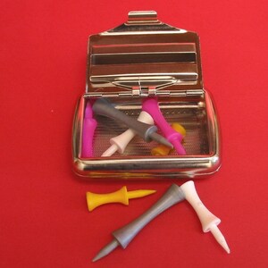 May include: A silver metal case with a hinged lid, containing a collection of colorful golf tees. The case is open, revealing the tees inside. There are also three golf tees lying outside the case on a red surface.