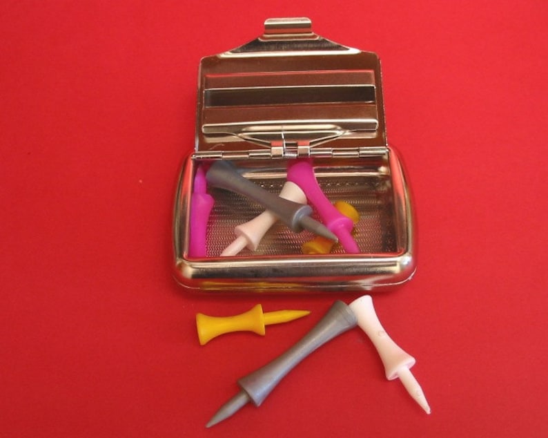 May include: A silver metal case with a hinged lid, containing a collection of colorful golf tees. The case is open, revealing the tees inside. There are also three golf tees lying outside the case on a red surface.