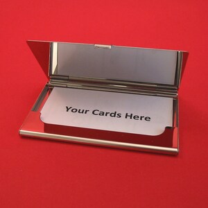 Puede incluir: Un porta tarjetas de metal plateado con una tarjeta blanca en el interior que dice "Your Cards Here".