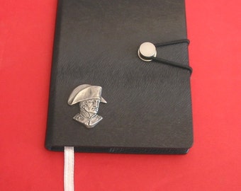 May include: A black leather-bound journal with a silver metal clasp and a silver metal portrait of a man in a hat. The journal has a white ribbon bookmark.