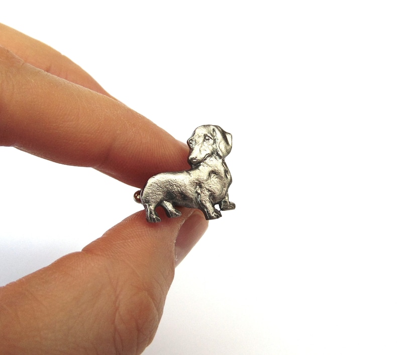 Puede incluir: Una figurita de dachshund de plata, una raza de perro popular, se muestra de perfil. La figurita es detallada y realista, con un acabado liso y met&aacute;lico.
