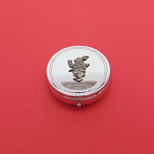 May include: A silver round pill box with a white rabbit design on the lid. The rabbit is wearing a waistcoat and is holding a pocket watch.