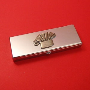 May include: A rectangular, silver business card holder with a decorative chef's hat emblem in gold and bronze tones. The card holder has a shiny, reflective surface and a small clasp. The background is a solid red.
