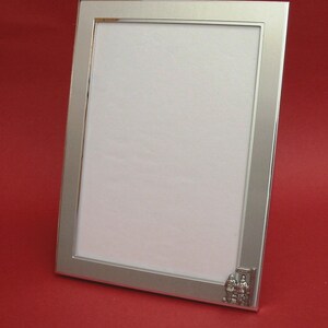 May include: Silver rectangular photo frame with a white interior. The frame has a simple design with a small decorative element in the lower right corner. The frame is set against a red background.