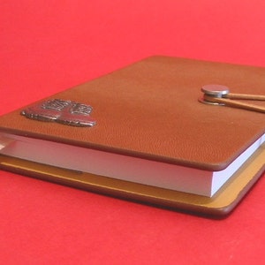 May include: A brown leather-bound journal with a metal boot charm and a ribbon closure. The journal is closed and lying on a red surface.