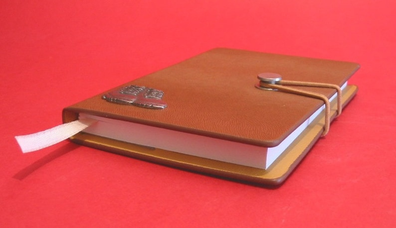 May include: A brown leather-bound journal with a metal boot charm and a ribbon closure. The journal is closed and lying on a red surface.