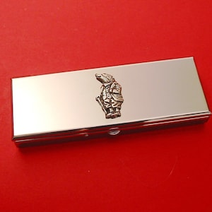 Könnte beinhalten: Eine silberne Pillendose aus Metall mit einem kleinen, silbernen 3D-Bild eines Mannes mit Hut und Spazierstock auf dem Deckel.
