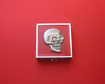 May include: A silver-colored skull emblem is centered on a red leather square, encased in a silver-toned metal frame. The skull has detailed features, including eye sockets and teeth. The item is set against a solid red background.