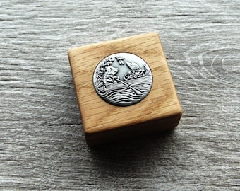 May include: A square wooden box featuring a circular silver-toned metal inlay on the lid. The inlay depicts a detailed relief of a small animal rowing a boat on water, with a dragonfly hovering above and another animal figure nearby.