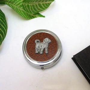 May include: A circular, silver-coloured pill box with a brown leather centre. A small, detailed silver dog figure is in the centre. The pill box is on a white surface, with green leaves in the background.