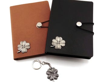 May include: Two rectangular notebooks, one brown and one black, each featuring a silver-toned four-leaf clover emblem and a circular button closure with an elastic band. A matching silver-toned four-leaf clover keychain is included.