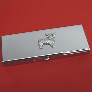 May include: A rectangular, silver business card holder with a Highland cow emblem. The card holder is set against a red background. The cow is detailed with textured fur and horns. The item is likely made of metal.