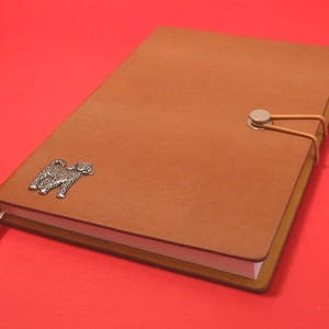 K&ouml;nnte beinhalten: Ein braunes Lederjournal mit einem silbernen Hundefolie und einem silbernen Verschluss. Das Journal hat einen wei&szlig;en Buchschnitt und ein braunes Lesezeichenband.