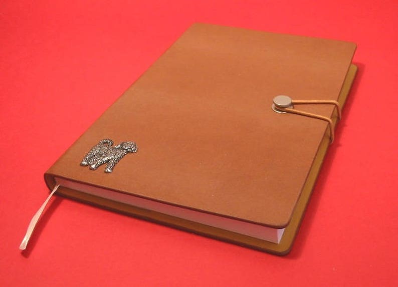 K&ouml;nnte beinhalten: Ein braunes Lederjournal mit einem silbernen Hundefolie und einem silbernen Verschluss. Das Journal hat einen wei&szlig;en Buchschnitt und ein braunes Lesezeichenband.