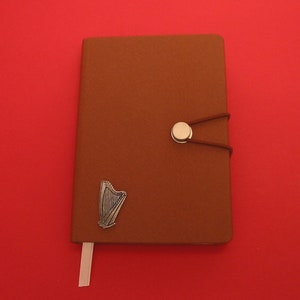May include: A brown leather-bound journal with a silver harp charm and a silver button closure. The journal has a white ribbon bookmark.