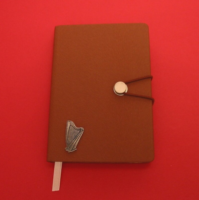 May include: A brown leather-bound journal with a silver harp charm and a silver button closure. The journal has a white ribbon bookmark.