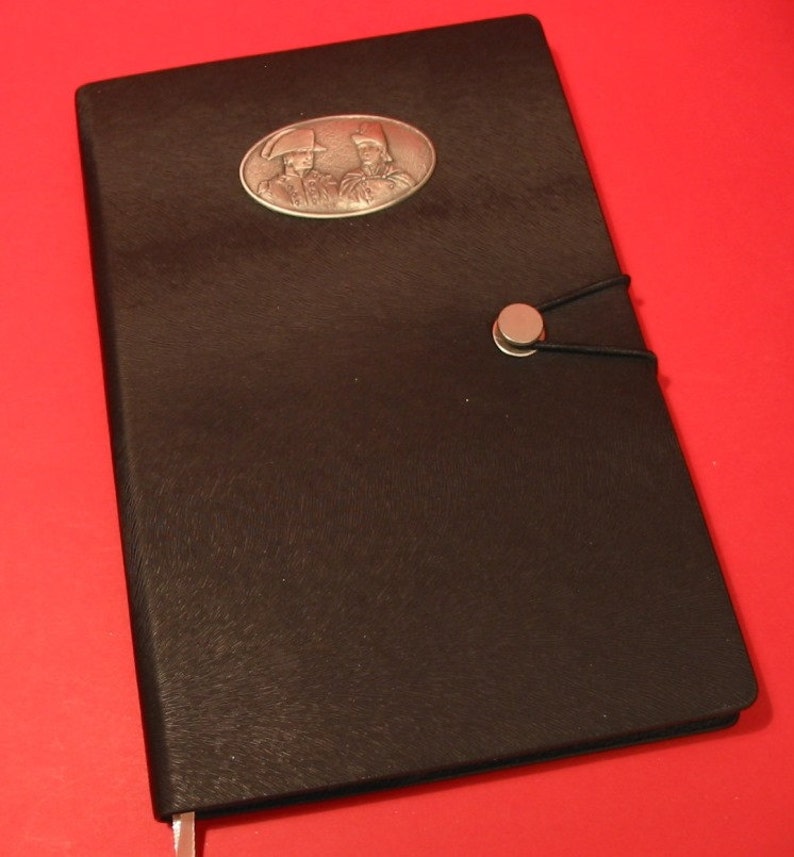 K&ouml;nnte beinhalten: Schwarzes Lederjournal mit einem ovalen Metall-Emblem, das zwei Figuren in historischer Kleidung zeigt. Das Journal hat einen silbernen Knopfverschluss mit einem schwarzen Gummiband. Ein silberner Lesezeichen ist sichtbar. Der Hintergrund ist rot.
