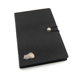 May include: Black leather journal with a silver cat emblem. The journal has a black elastic closure with a silver button. The rectangular journal has a textured surface. The cat emblem is on the lower left.