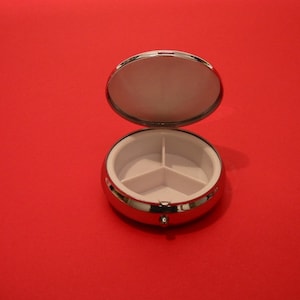 K&ouml;nnte beinhalten: Eine silberne Pillendose mit einem Scharnierdeckel und drei F&auml;chern. Der Deckel ist ge&ouml;ffnet und zeigt die leeren F&auml;cher.